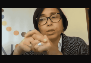 Persona con gafas y cabello corto, vistiendo un suéter a rayas, con las manos unidas frente al rostro.