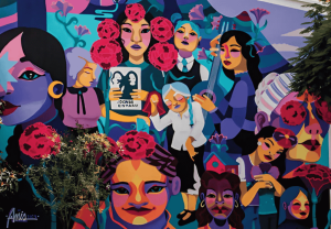Un vibrante mural muestra a mujeres diversas ataviadas con ropas coloridas y rodeadas de elementos florales. Algunas sostienen libros, mientras que otras realizan diversas actividades, todo ello sobre un fondo abstracto y animado. Un árbol oculta parcialmente el cautivador diseño del mural.