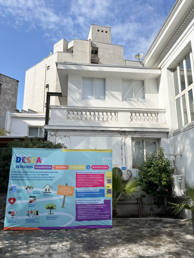 Un cartel informativo sobre los derechos económicos, sociales, culturales y ambientales (DESCA) está situado frente a un edificio de color blanco. El cartel tiene un diseño colorido con secciones de información y códigos QR, mientras que el cielo azul y nubes blancas son visibles en el fondo.