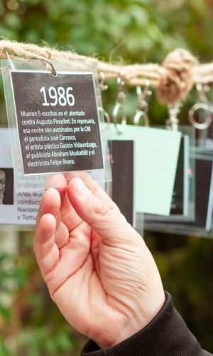 Mano sosteniendo una placa de plástico transparente con texto que dice "1986, mueren 5 escoltas en el atentado contra Augusto Pinochet. En represalia, esa noche son asesinados por la CNI el periodista José Carrasco, el artista plástico Gastón Vidaurrázaga, el publicista Abraham Muskatblit y el electricista Felipe Rivera.” La placa está colgada de una cuerda con otras similares y el fondo es borroso mostrando vegetación.