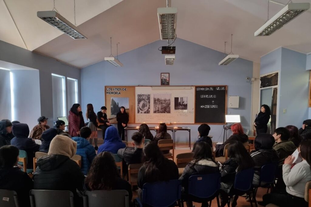Personas reunidas en una sala con carteles y velas sobre una mesa en el frente, en un evento titulado "Los sitios de memoria".