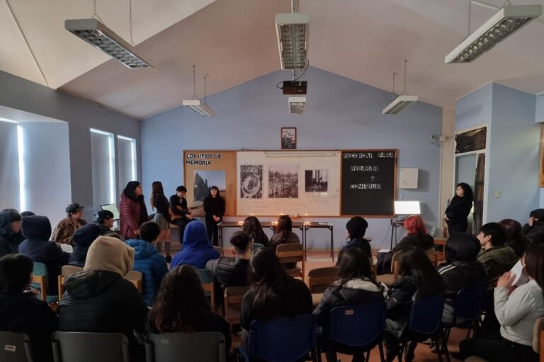 Personas reunidas en una sala con carteles y velas sobre una mesa en el frente, en un evento titulado "Los sitios de memoria".