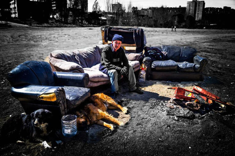Hombre sentado en un sofá al aire libre junto a dos perros, con edificios al fondo.