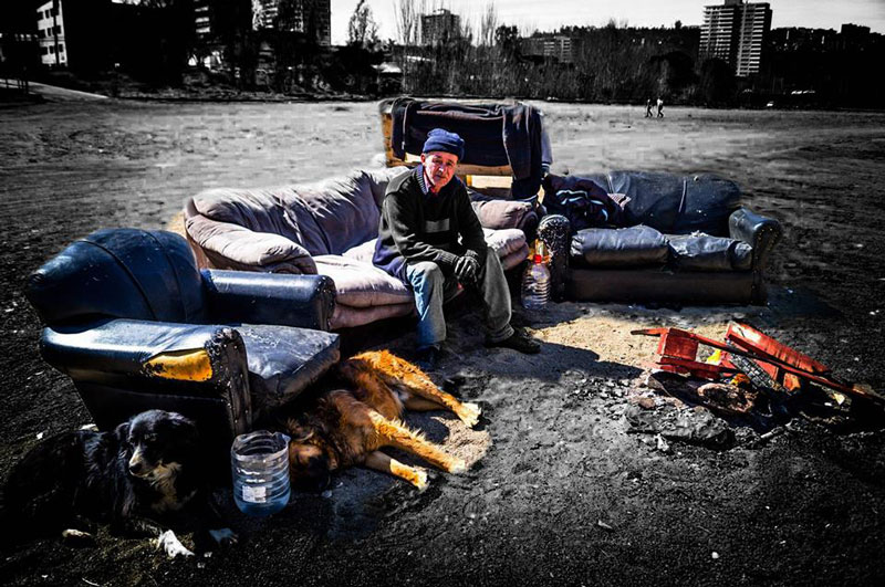 Hombre sentado en un sofá al aire libre junto a dos perros, con edificios al fondo.