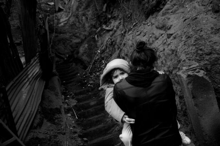 Mujer vestida de oscuro cargando a un niño abrigado, subiendo una escalera de tierra rodeada de paredes de tierra y metal.