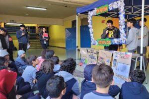 Grupo de estudiantes sentados en un aula, escuchando a un hombre hablar bajo un cartel que dice "Memoria Rebelde", con carteles educativos alrededor.