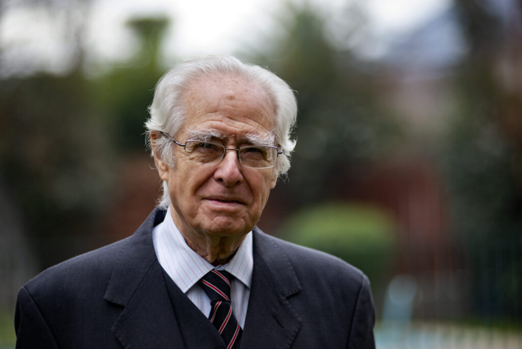 Hombre mayor con traje oscuro y corbata en un entorno al aire libre, con fondo desenfocado.