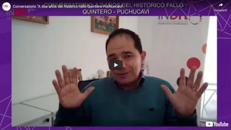 Una persona está sentada en una oficina, gesticulando con ambas manos en alto. Detrás de ella hay una pancarta con el logo del INDH y un texto sobre un fallo histórico de Quintero-Puchuncaví. El escenario parece ser un conversatorio o entrevista virtual.