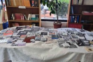 Mesa cubierta con libros y documentos, rodeada de estanterías llenas de libros y una planta en maceta al fondo.