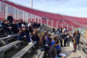 Grupo de estudiantes sentados en una gradería de estadio, escuchando a una persona de pie.