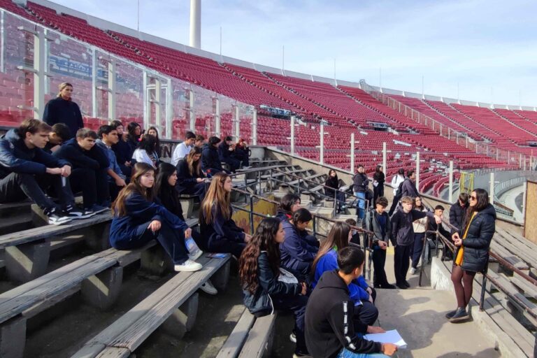 Grupo de estudiantes sentados en una gradería de estadio, escuchando a una persona de pie.