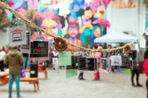 Instalación artística con tarjetas colgando de una cuerda, personas al fondo y un mural colorido en la pared.