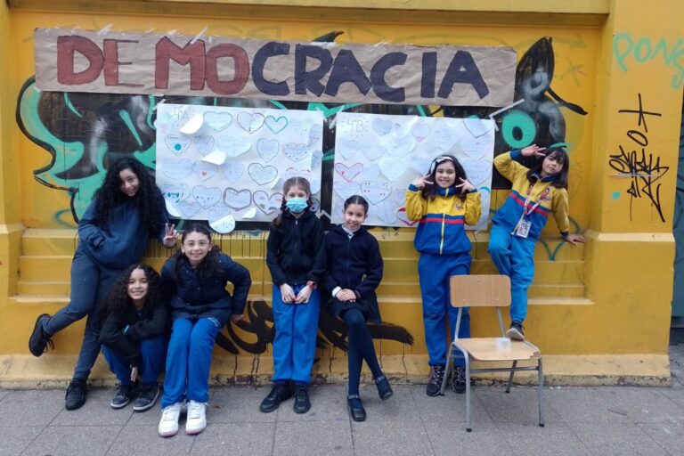Grupo de niños frente a un mural de "DEMOCRACIA" con corazones pintados, posando frente a una pared amarilla con graffiti.