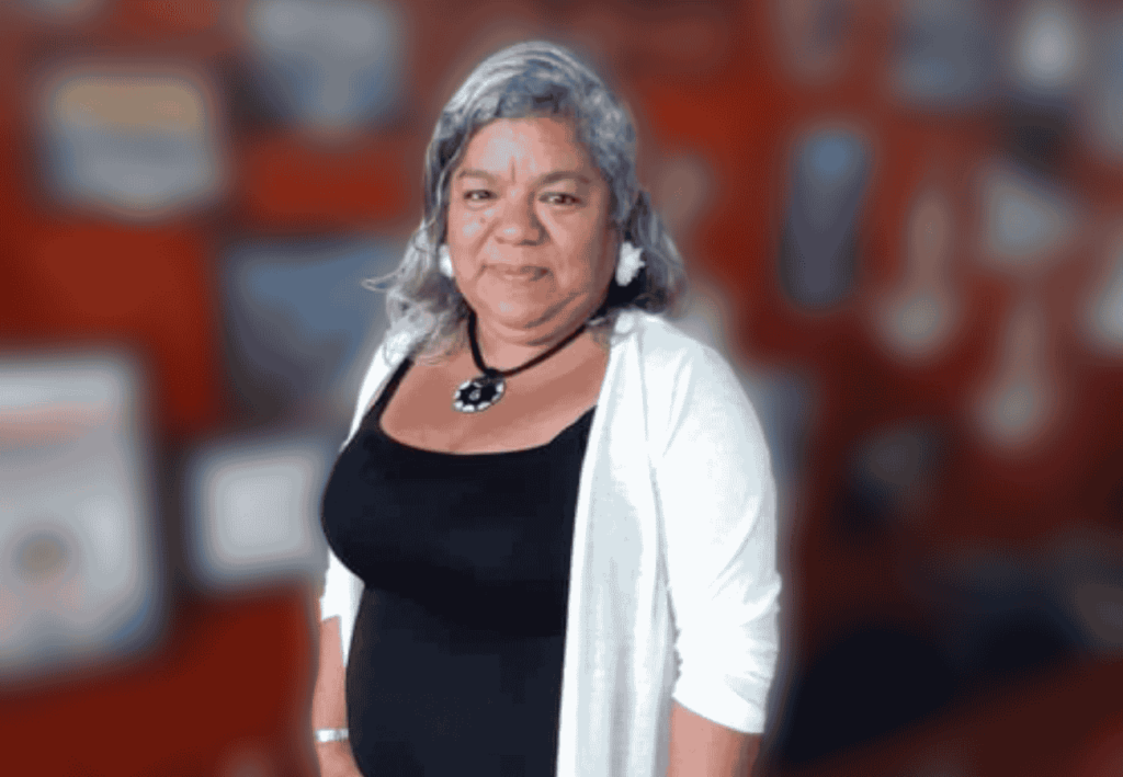 Mujer de pie con cabello canoso, vestida de negro y chaqueta blanca, fondo desenfocado de color rojo.