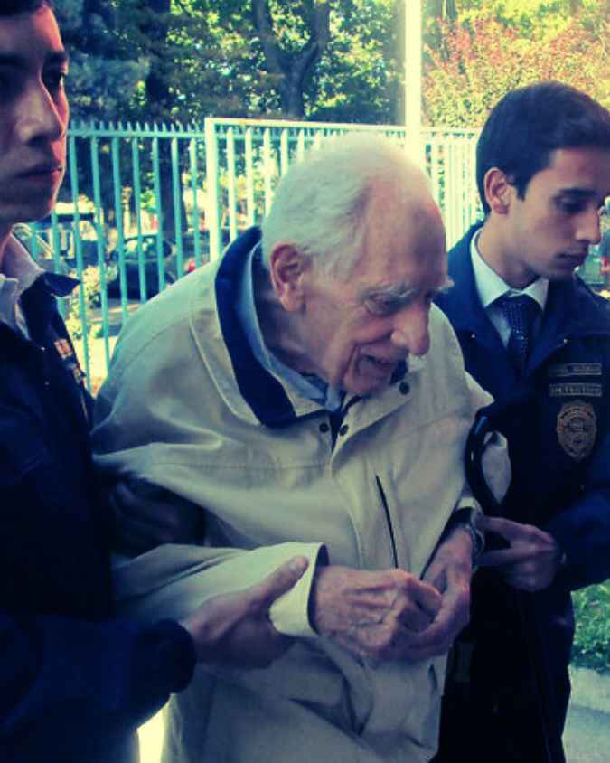 Un hombre anciano con abrigo beige es asistido por dos personas que parecen ser oficiales de policía, mientras camina al aire libre con árboles y una cerca metálica en el fondo.