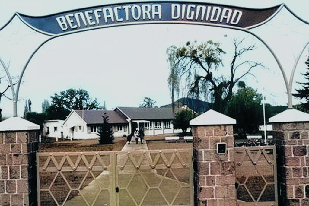 Entrada de un complejo con un letrero que dice 'Benefactora Dignidad', mostrando un camino hacia un edificio de estilo simple al fondo, rodeado de árboles.