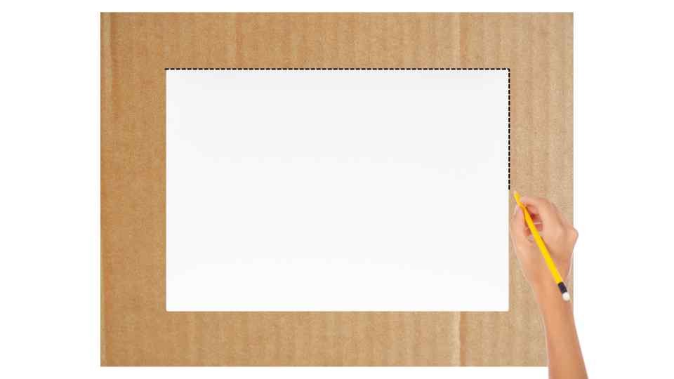 Una mano sosteniendo un lápiz marca el borde de un rectángulo blanco sobre un fondo de cartón marrón claro.