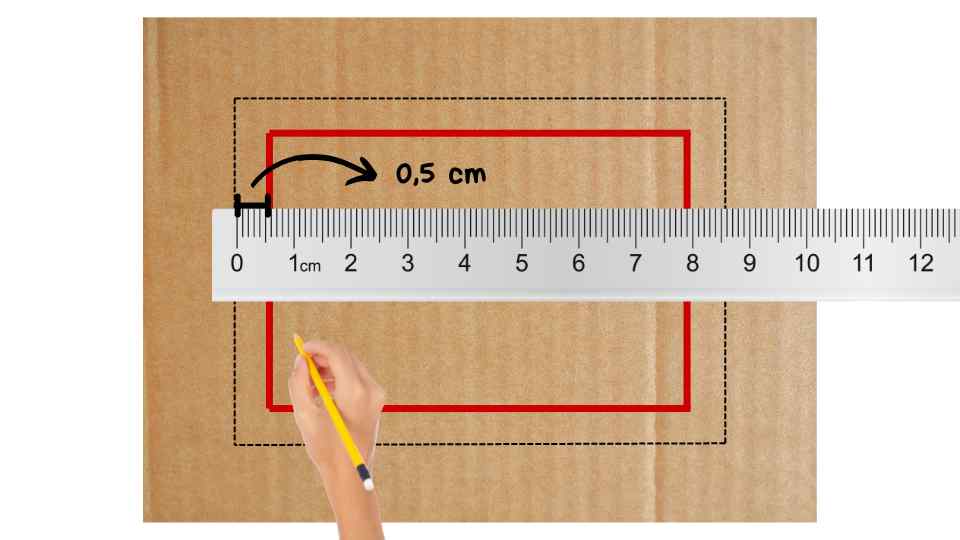 Una regla mide un espacio de 0,5 cm entre un rectángulo negro punteado y uno rojo sólido sobre una superficie de cartón, mientras una mano sostiene un lápiz amarillo.