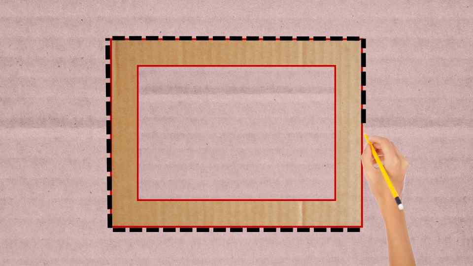 Una mano sostiene un lápiz amarillo y traza una línea en un rectángulo de cartón con un borde negro y rojo, sobre un fondo rosa claro.