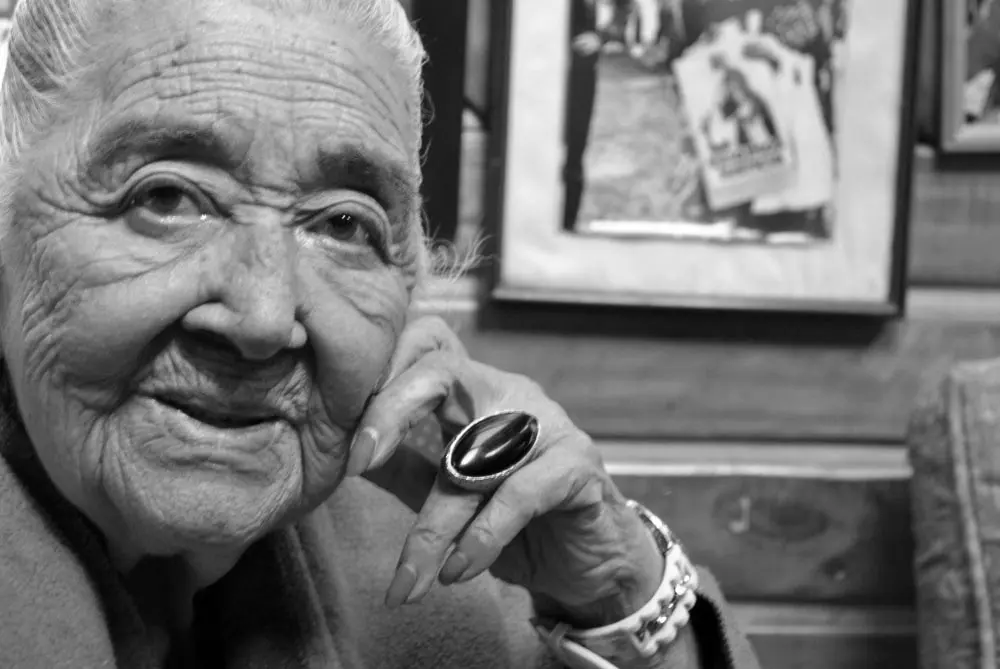 Fotografía en blanco y negro de una mujer mayor con una expresión serena, vistiendo un abrigo y un anillo grande y oscuro en su dedo índice. Tiene el cabello canoso recogido y muestra una sonrisa leve. De fondo, hay cuadros colgados en la pared.