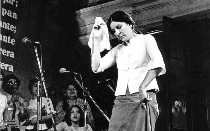 Mujer joven vestida con blusa blanca y falda larga bailando en un escenario, levantando una mano que sostiene un pañuelo. Detrás de ella, hay un grupo de personas con micrófonos, posiblemente cantando o tocando música. El ambiente es de una presentación en vivo.