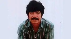 Hombre con cabello oscuro y bigote, vistiendo una camisa a rayas verdes, posando frente a un fondo blanco.