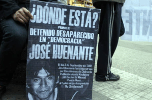 Cartel en la calle con la imagen de una persona desaparecida, preguntando "¿Dónde está?" y destacando a José Huenante como el primer detenido desaparecido en democracia en Chile. El texto se refiere a su secuestro en septiembre de 2005 por Carabineros en Puerto Montt y enfatiza la importancia de no olvidar su desaparición. Una persona sostiene el cartel mientras está sentada en una acera.
