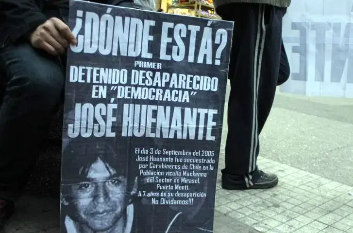 Persona sosteniendo un cartel que pregunta "¿Dónde está? Primer detenido desaparecido en 'democracia' José Huenante", con una foto y detalles sobre su desaparición en 2005 en Puerto Montt, Chile.