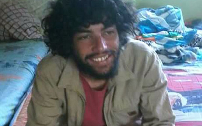 Persona con cabello rizado y barba sonríe, sentado en una habitación con una cama detrás de él.