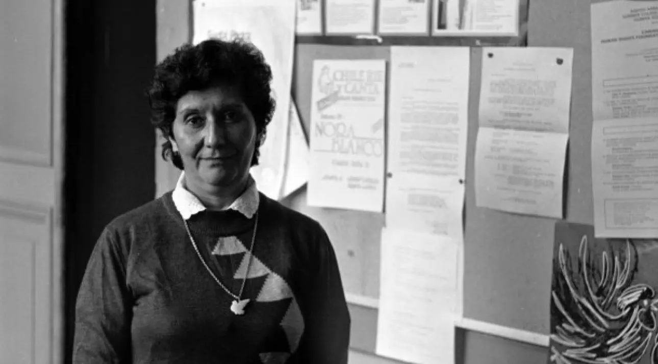 Mujer de pie frente a un tablero con papeles y anuncios en la pared, en una fotografía en blanco y negro.