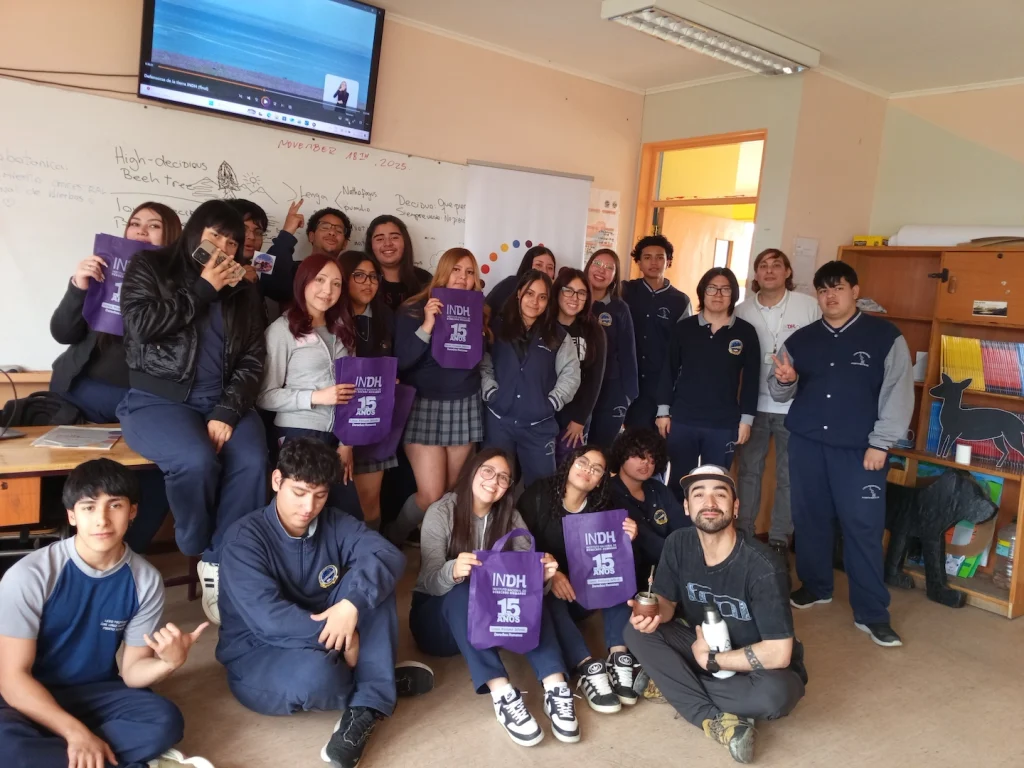 Grupo de estudiantes posando en un aula, algunos sosteniendo bolsas moradas con el texto "INDH 15 años".