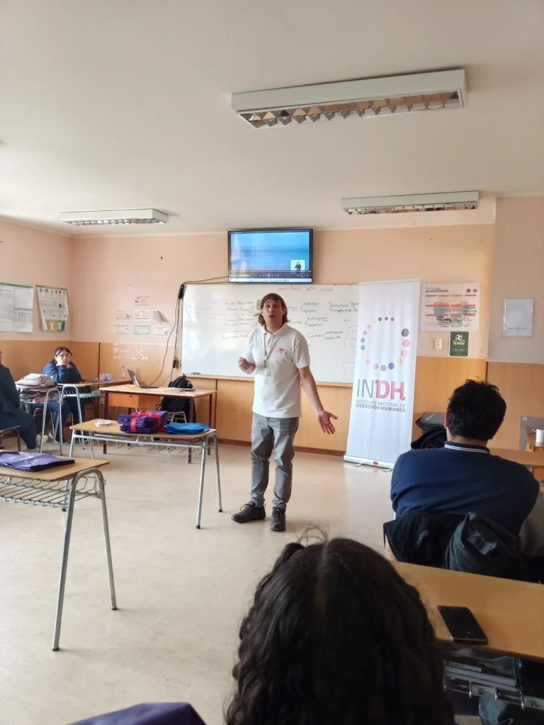Persona de pie hablando frente a una clase, con una pantalla y un cartel del INDH en el fondo.