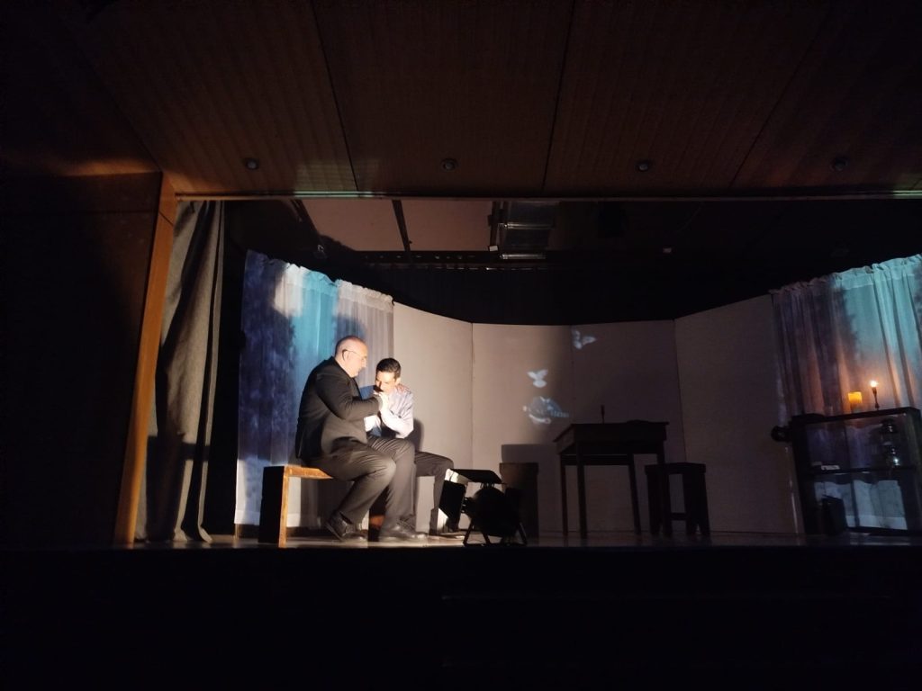 Dos actores en un escenario sentado en un banco de madera, iluminados por un foco, en lo que parece ser una representación teatral. El fondo del escenario tiene cortinas blancas y una mesa pequeña con una vela encendida.