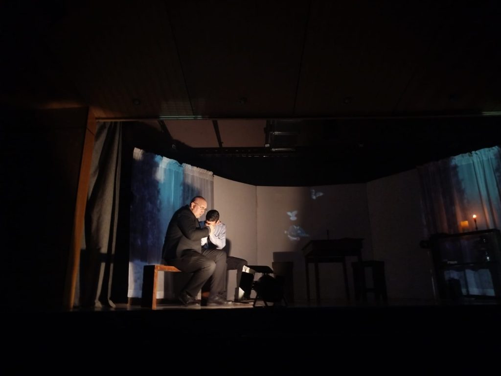 Dos personas sentadas en un banco en el escenario de un teatro, con luces tenues y una vela encendida al fondo; una parece reconfortar a la otra mientras mantienen la cabeza baja.