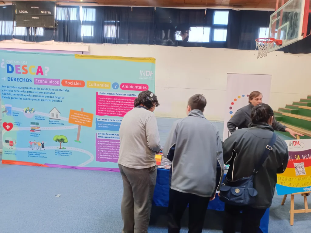 Personas interactuando en un stand del Instituto Nacional de Derechos Humanos con un cartel sobre derechos económicos, sociales, culturales y ambientales en un gimnasio.