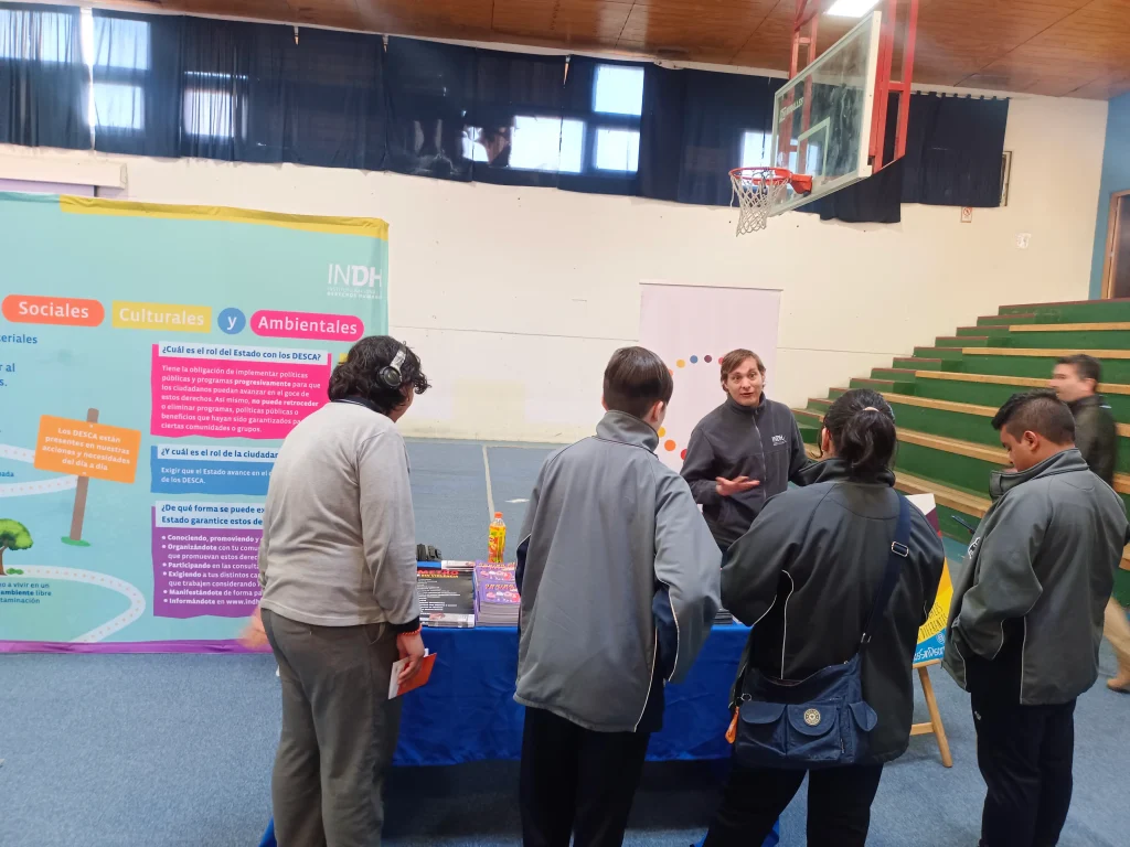 Personas reunidas alrededor de un stand informativo en un gimnasio, donde un hombre explica algo a varios jóvenes; al fondo se ve un cartel del INDH sobre derechos sociales, culturales y ambientales.