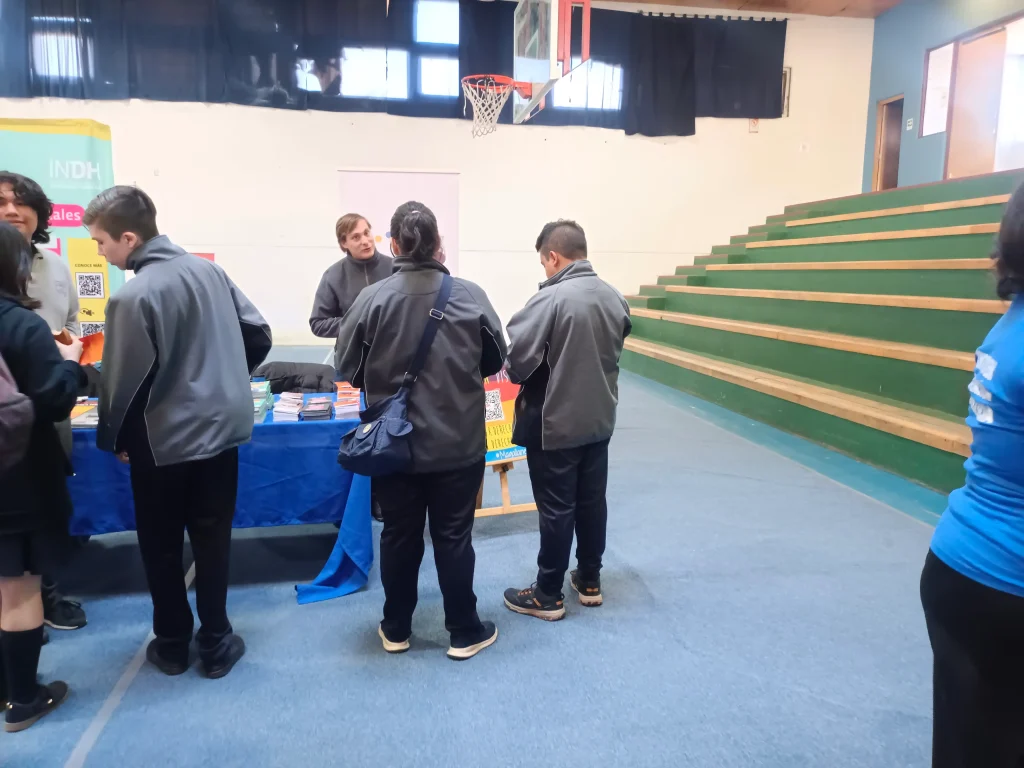 Personas en una feria escolar observando una mesa con folletos, ubicada en un gimnasio con graderías vacías y cortinas negras en el fondo.