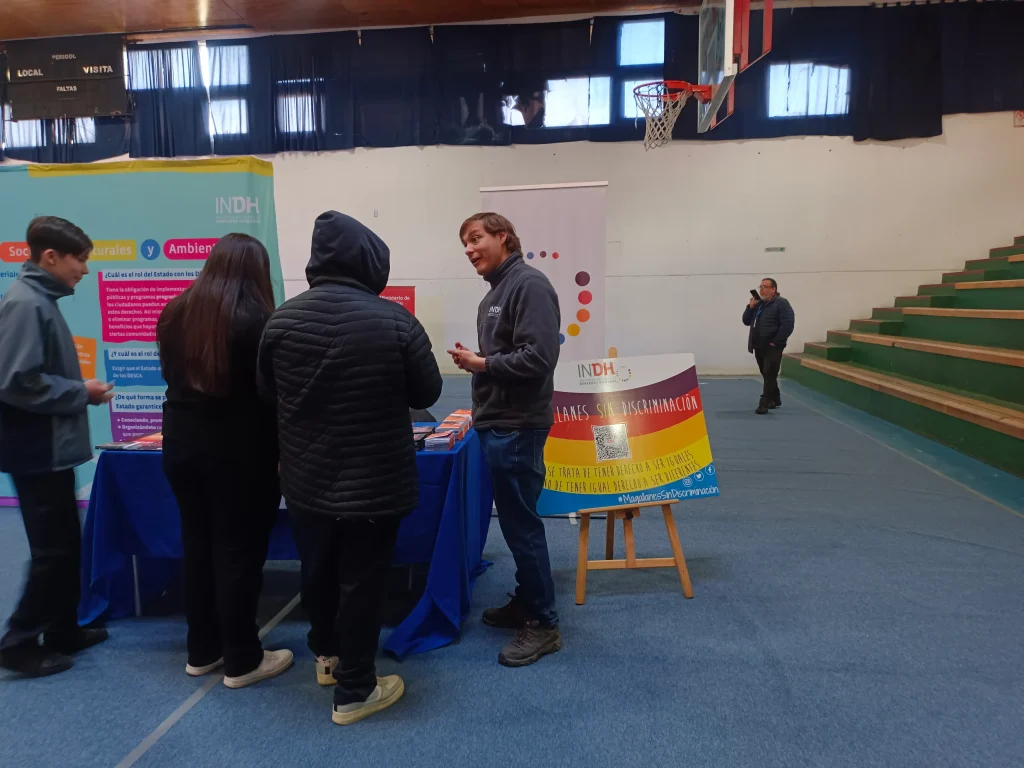 Varias personas conversan en un stand informativo del INDH en un gimnasio. Hay carteles sobre derechos sociales, culturales y sin discriminación.