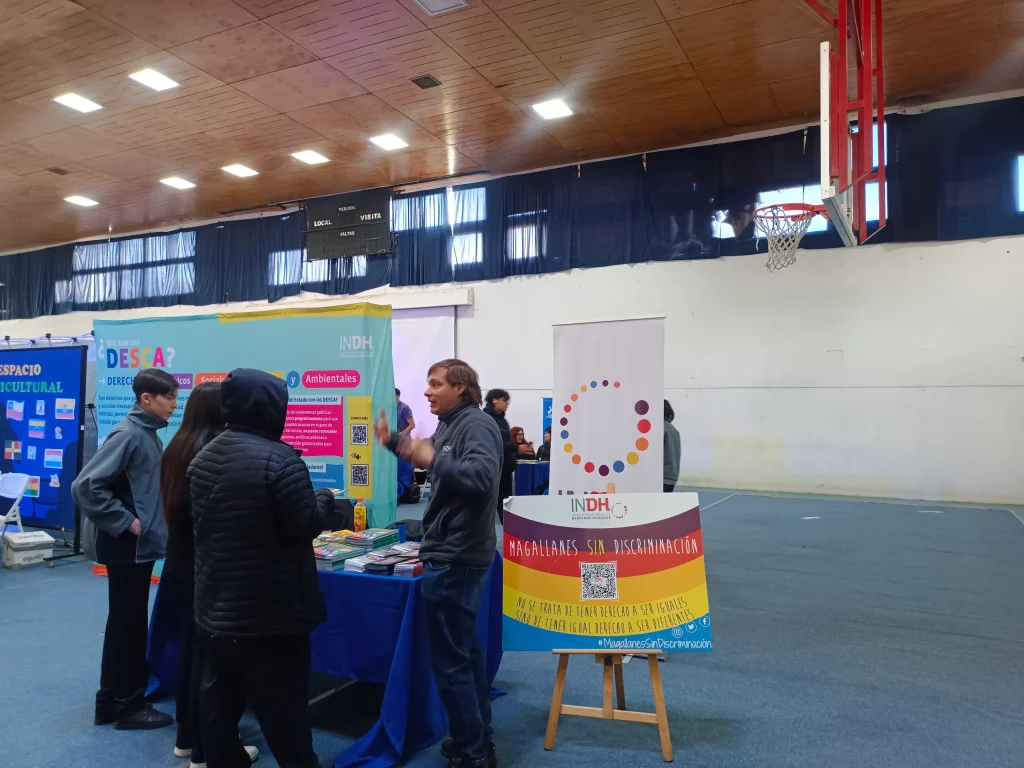 Personas conversando en un stand informativo en un polideportivo, con carteles sobre derechos y la frase 'Magallanes sin discriminación'.