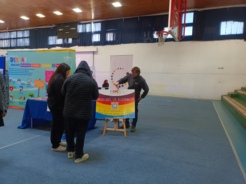 Personas interactúan en un evento informativo en un gimnasio, con un cartel de "Magallanes sin discriminación" en un caballete y un fondo colorido sobre derechos económicos y sociales.