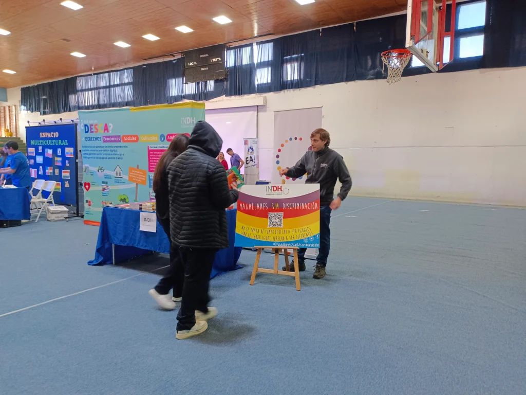 Un evento en un gimnasio donde varias personas interactúan cerca de puestos informativos sobre derechos humanos y no discriminación. En el fondo hay paneles con información y carteles relacionados con derechos económicos y sociales.