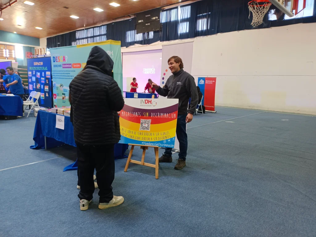 Una persona de pie junto a un cartel del Instituto Nacional de Derechos Humanos (INDH) que dice "Magallanes sin discriminación" en un gimnasio durante un evento de información con mesas y carteles.