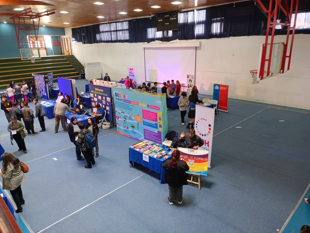 Feria informativa en un gimnasio con puestos de diferentes organizaciones, donde varias personas interactúan y se informan sobre diversos temas. Se observan mesas con folletos y carteles coloridos, y las gradas están al fondo a la izquierda.