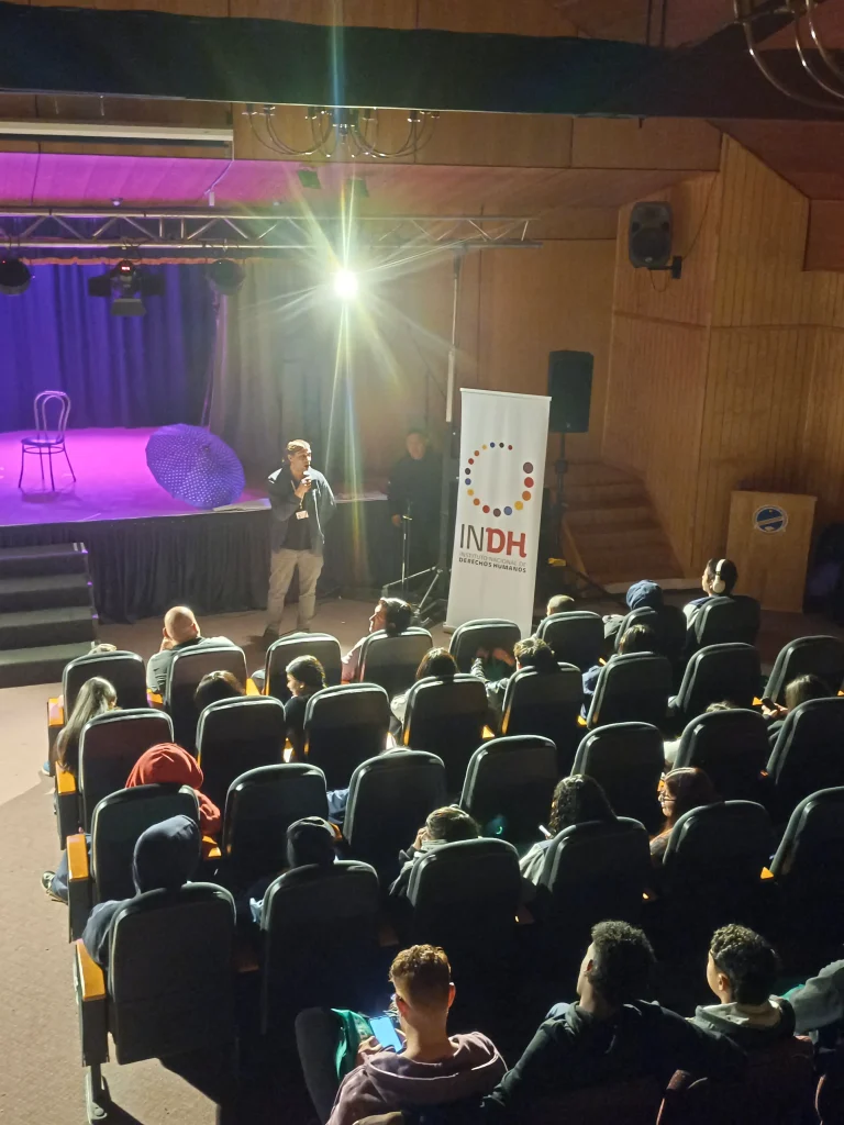 Una persona hablando en un escenario frente a una audiencia sentada en una sala, con un cartel del INDH al lado. Detrás de él, un escenario iluminado con una silla y un paraguas como decoración.