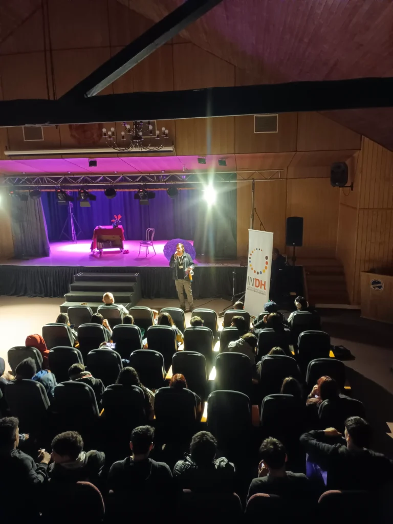 Auditorio con personas sentadas mirando hacia un escenario iluminado con luces de colores. En el escenario, hay una persona de pie junto a un micrófono y una pancarta con el logo "INDH". Se observa un fondo con cortinas azules y una mesa cubierta con un paño.