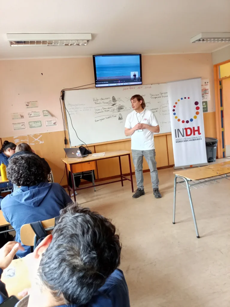 Un hombre de pie al frente de un aula, dando una presentación. Hay una pizarra con escritura y dibujos detrás de él, junto con un proyector que muestra una imagen de un paisaje costero en una pantalla. Varias personas están sentadas, prestando atención. A la derecha, un cartel con el logo del Instituto Nacional de Derechos Humanos.