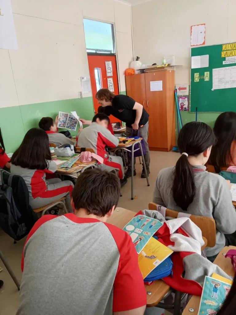 Niños sentados en un aula escolar vistiendo uniformes grises con detalles rojos mientras un adulto se inclina sobre uno de los estudiantes mostrando una hoja de ejercicios. El aula tiene paredes verdes y blancas, un armario de madera, y un tablón verde con papeles y dibujos pegados.