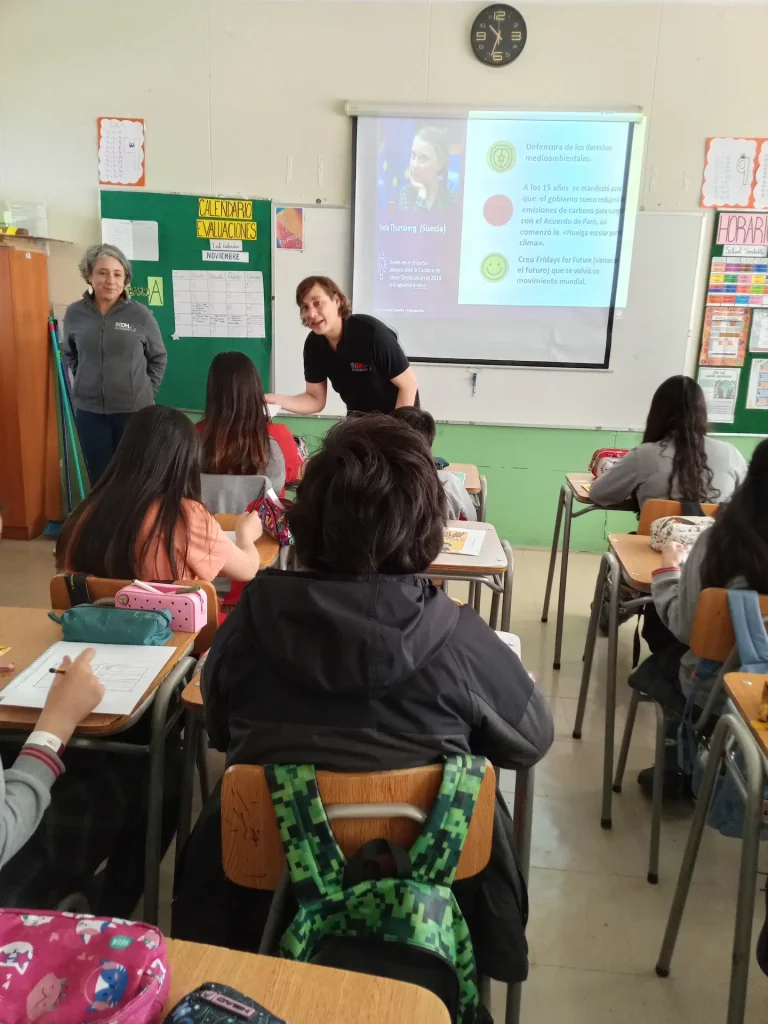 Clase en un aula escolar donde una persona está presentando información sobre Greta Thunberg y el cambio climático a un grupo de estudiantes sentados en sus escritorios. Hay un proyector que muestra una diapositiva con la imagen de Thunberg y texto sobre sus actividades medioambientales.