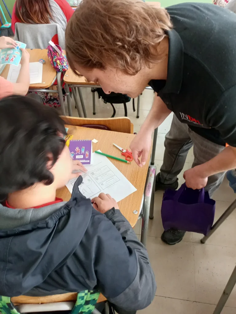 Un adulto ayuda a un niño con una actividad en un aula. El niño está sentado en un pupitre, trabajando con hojas de papel y tijeras. El adulto inclina su cuerpo hacia el niño mientras sostiene una bolsa morada en la mano. Al fondo, hay más niños sentados en sus pupitres.