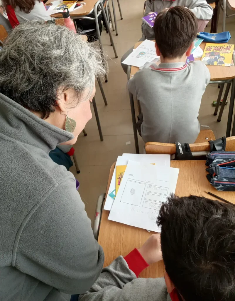 Una persona mayor revisa el trabajo de un estudiante en un aula, mostrando una hoja de ejercicios sobre una mesa junto a útiles escolares, mientras ambos están sentados.